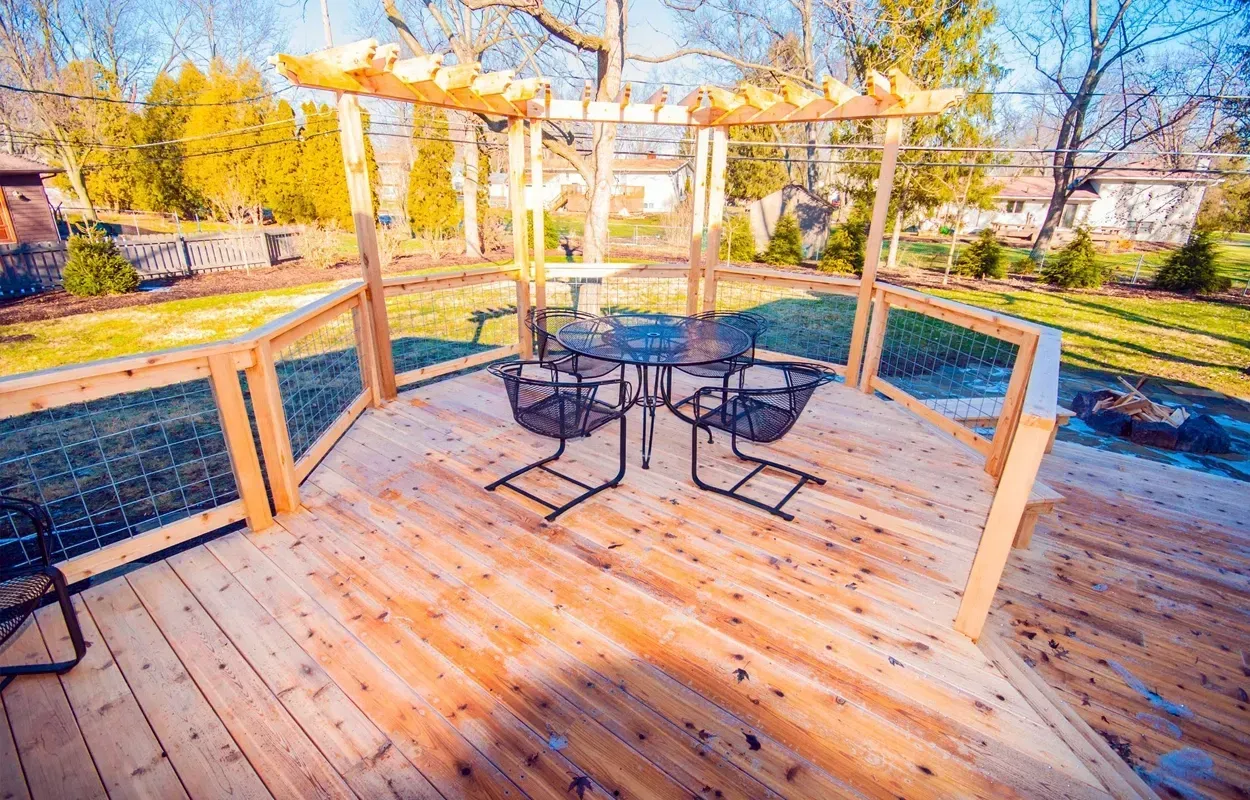 Wooden deck with black patio table and chairs, surrounded by railings and sunlit trees.