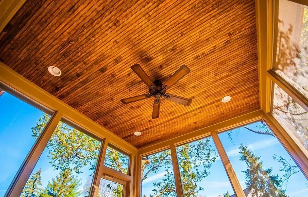 Wooden screened porch ceiling with fan, surrounded by windows and blue sky beyond.