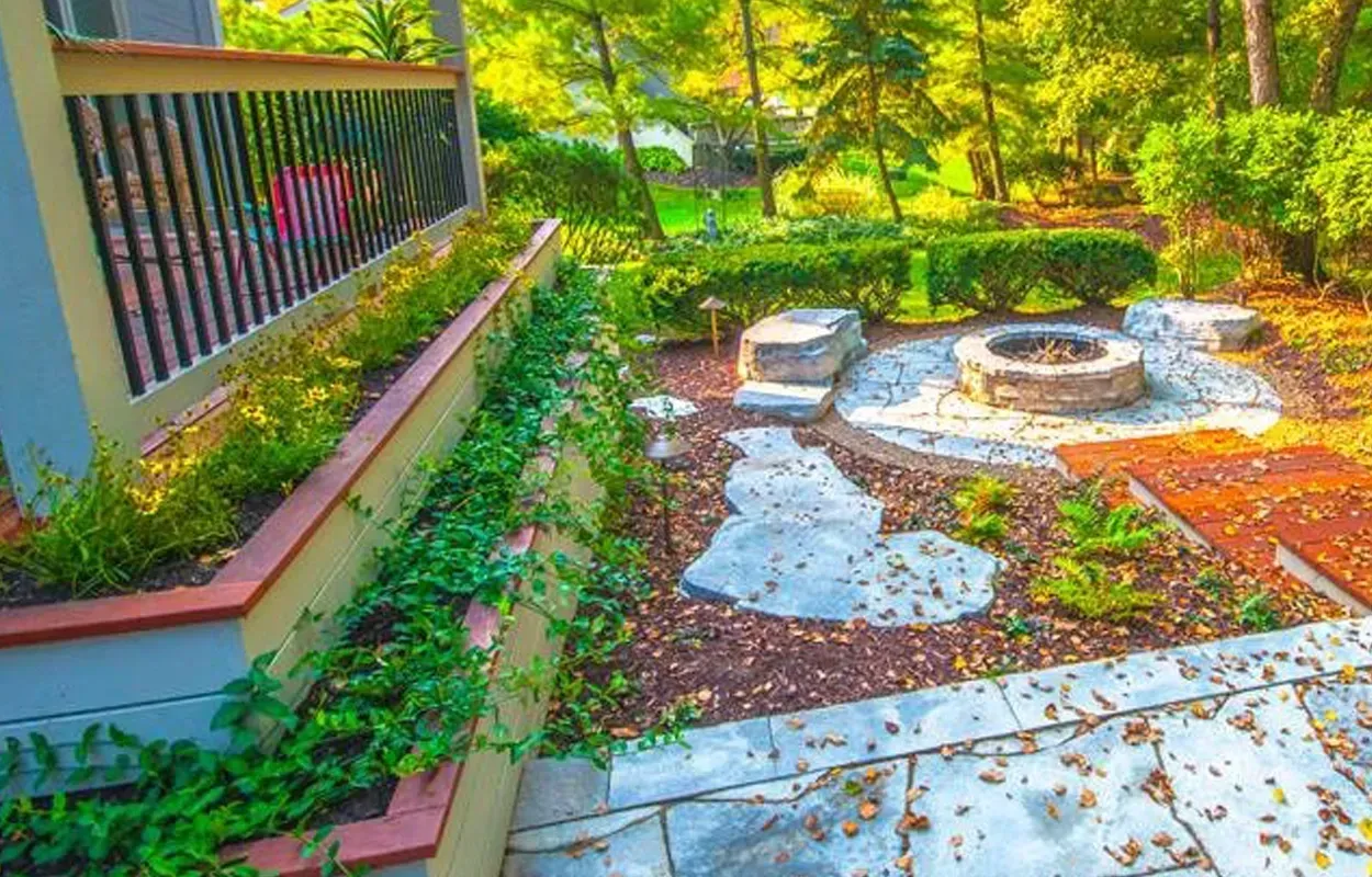 Sunny garden patio with stone path, circular fire pit, and lush green plants behind a wooden railing