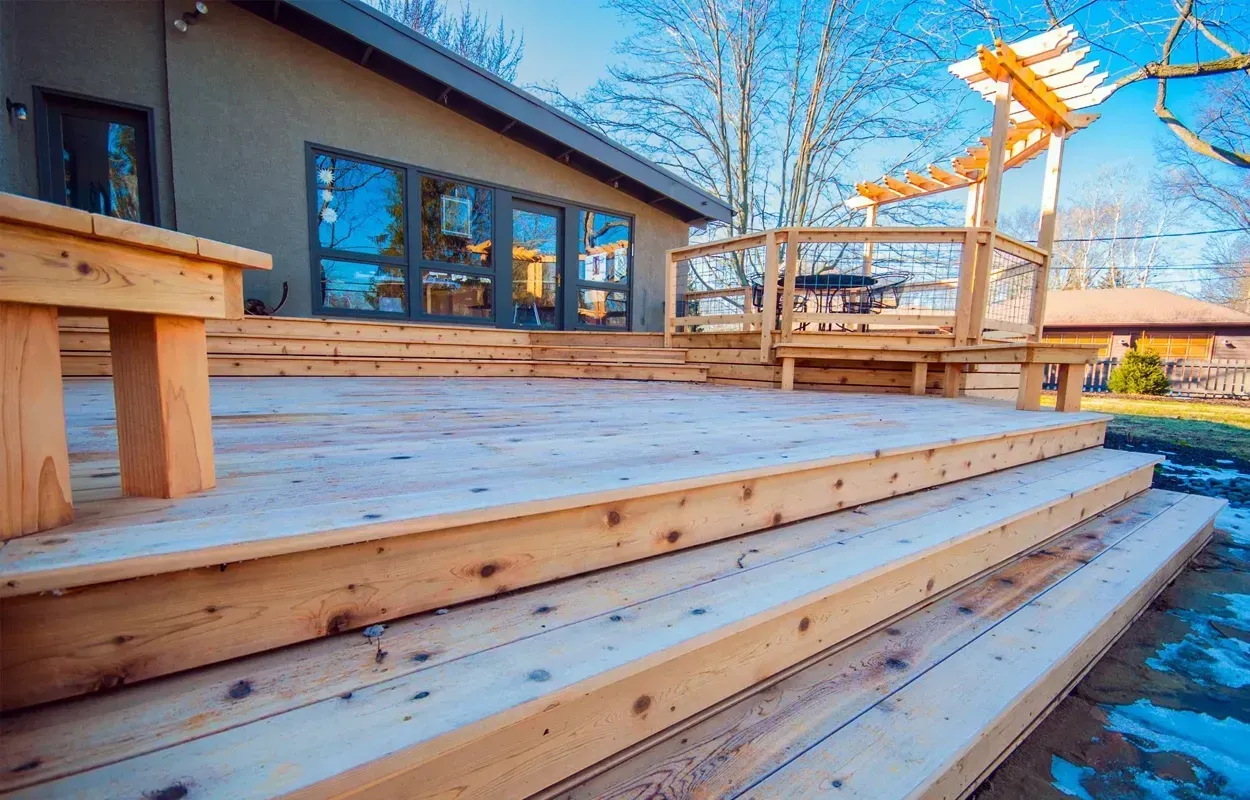 Wooden deck steps beside a house with windows, a pergola, and snowy ground in winter light