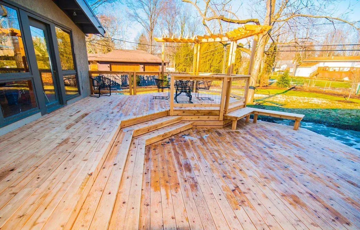 Wooden deck with steps and railing beside a house, overlooking a sunny backyard and playground swings.