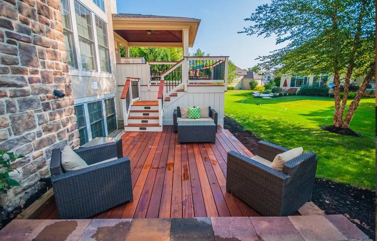Stone patio with wicker chairs, wooden deck stairs, and a green lawn beside a house