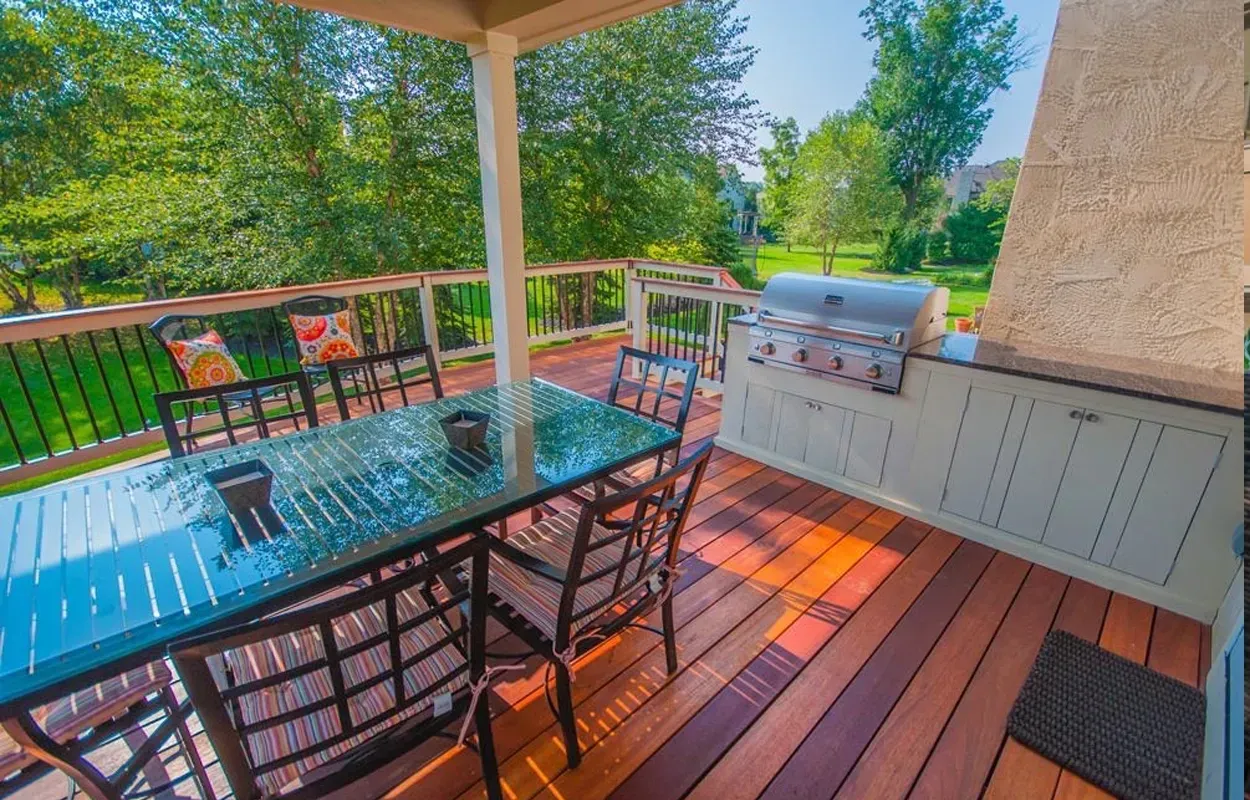 Covered deck with glass-top table, metal chairs, grill, and cabinets overlooking a green yard