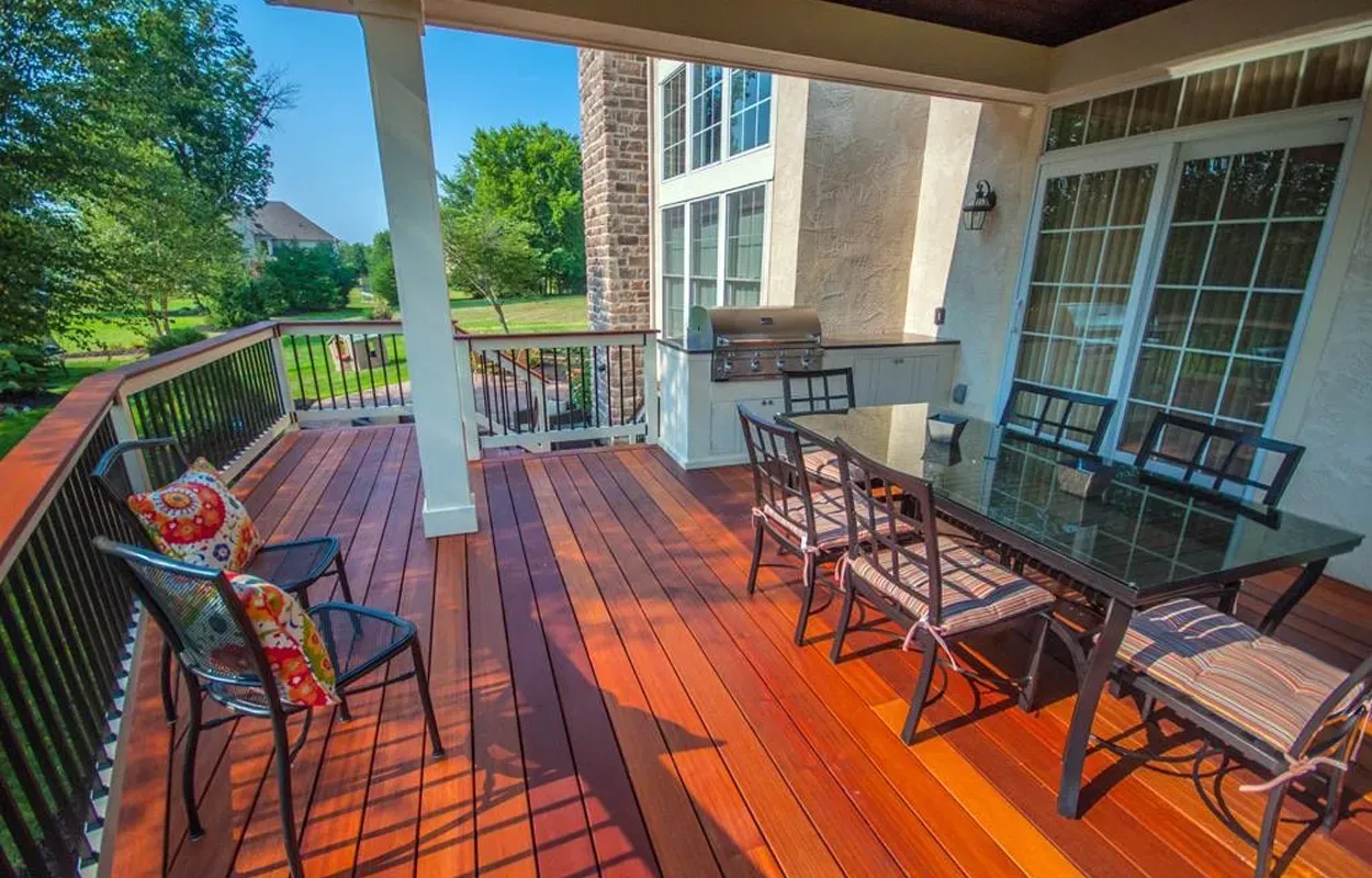 Covered wooden porch with glass dining table, chairs, and a railing overlooking greenery
