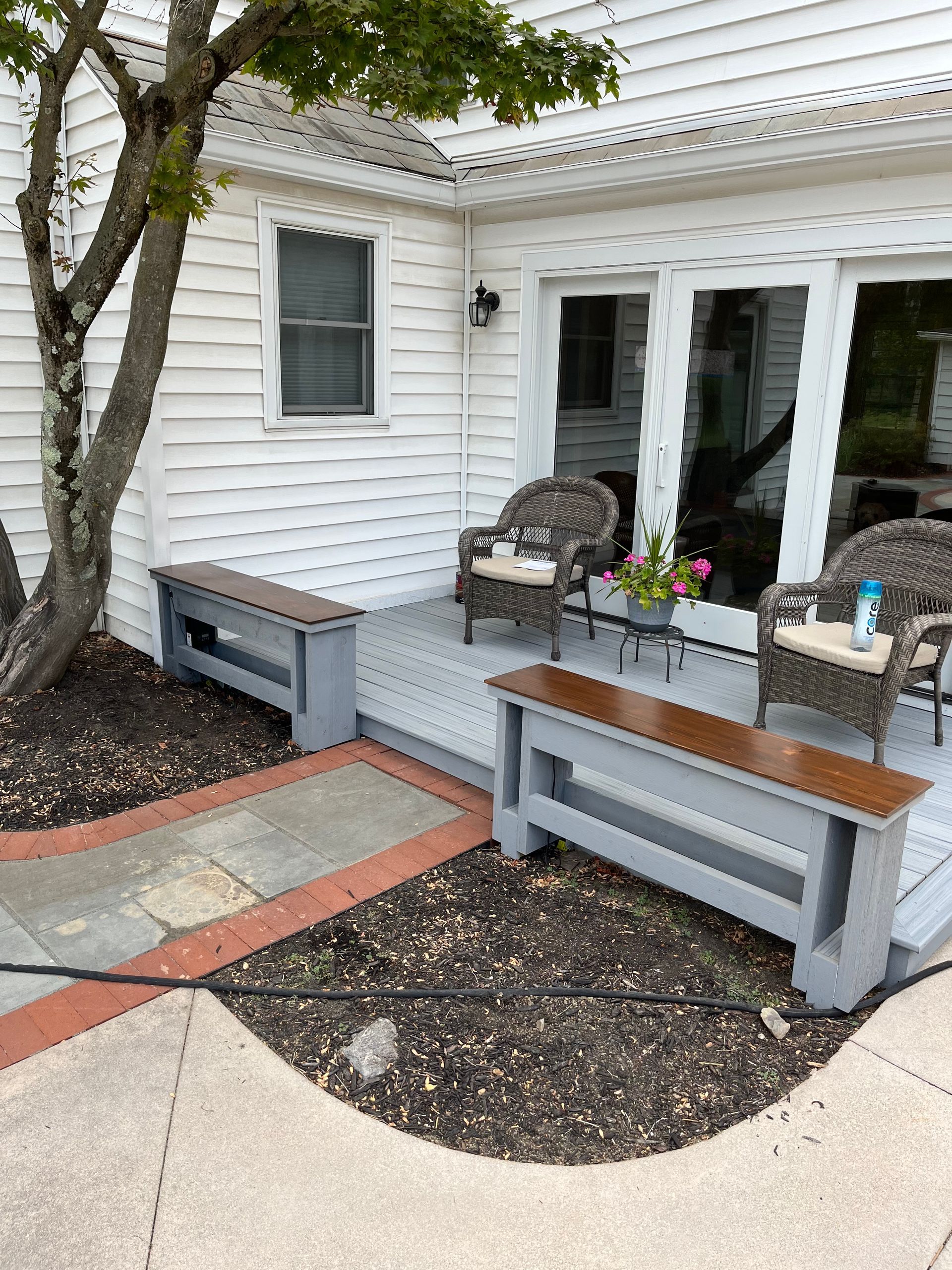 Front porch seating area with gray benches, wicker chairs, and a small table beside a white house.