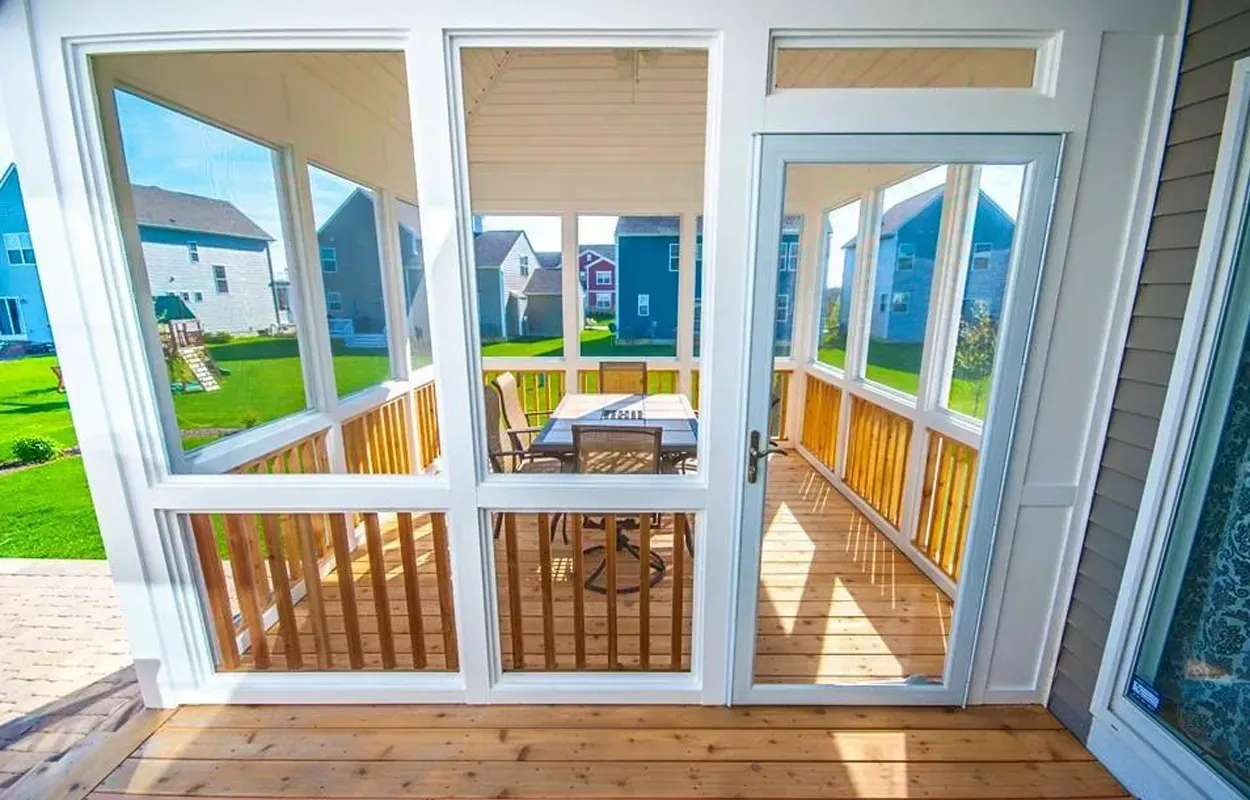 Enclosed porch with white-framed windows, wood flooring, and a small table, overlooking a green yard.