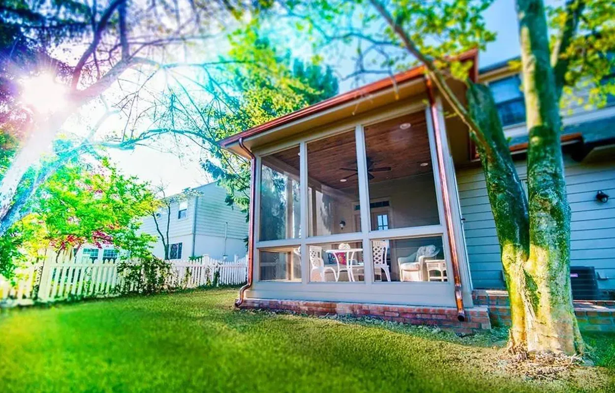 Sunny backyard with a covered wooden porch, green lawn, and trees beside a house