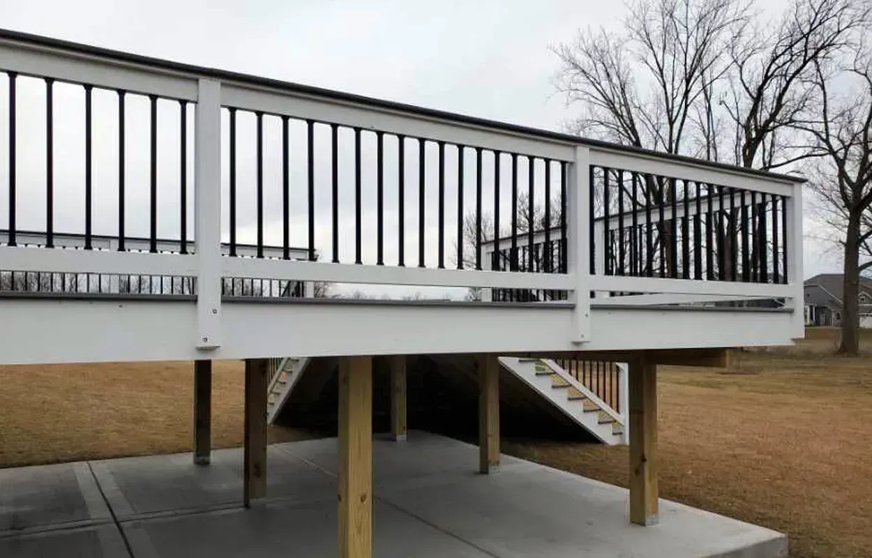 Elevated deck with black railing on wooden posts over a concrete patio in a bare yard