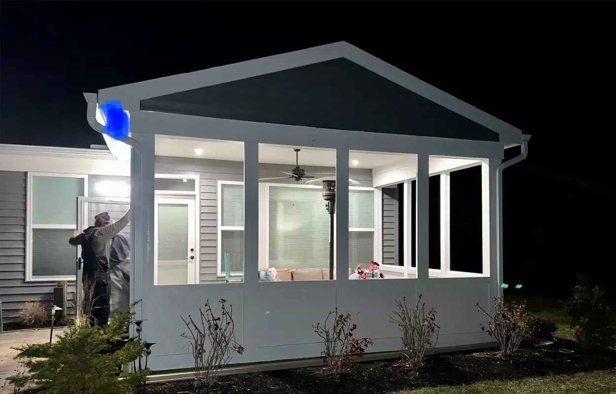 Person standing on a lit porch at night, painting a white house exterior