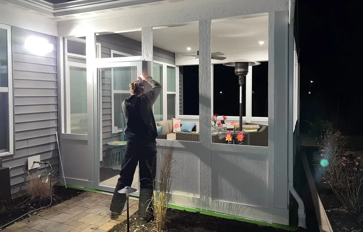 Person standing on a porch at night, reaching up near the ceiling under bright lights.