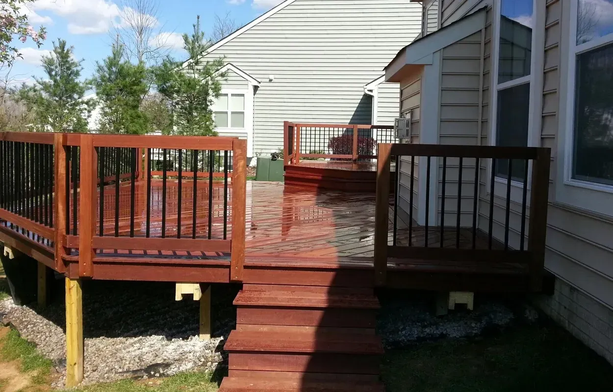 Wooden deck with red railings and steps beside a white house in sunlight