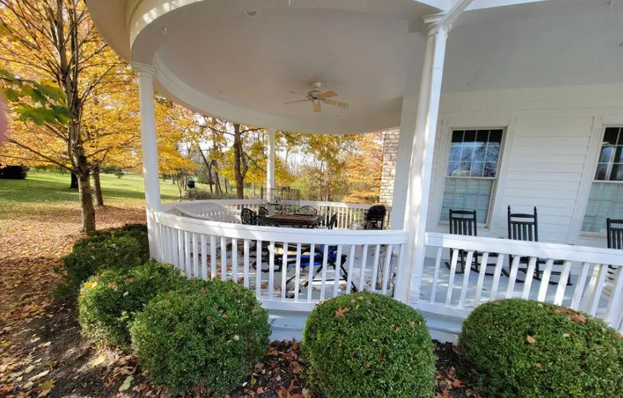 White porch with railing and chairs under a large curved roof, bordered by trimmed bushes and autumn trees.