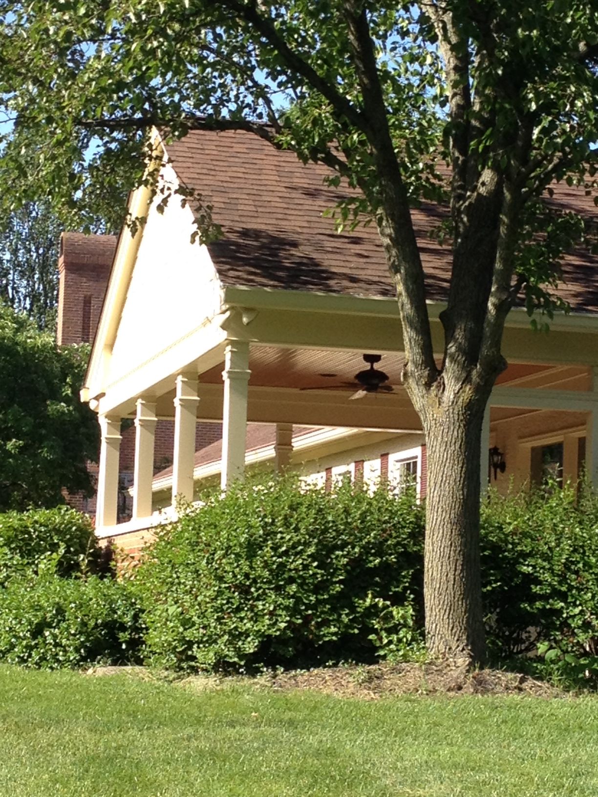 A house with a covered porch, white columns, and a large tree in front, surrounded by green lawn and bushes