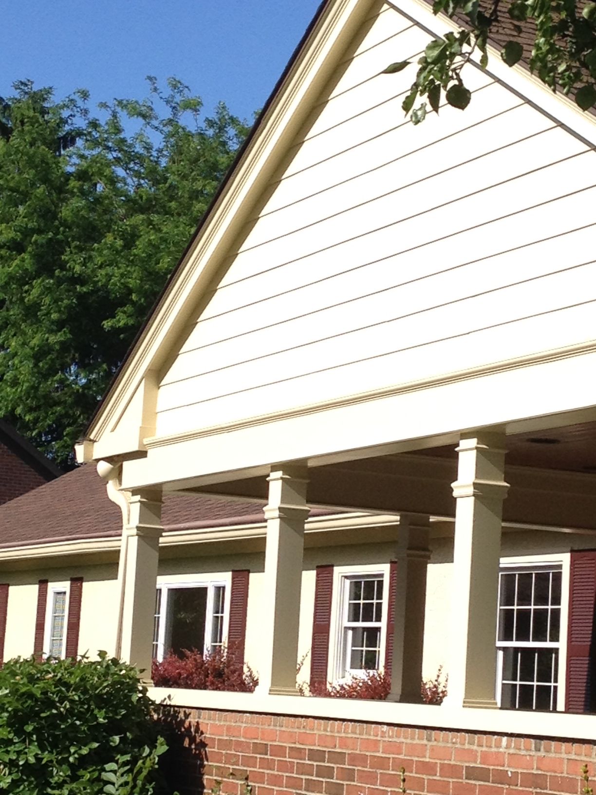 Front of a beige house with white trim, porch columns, and maroon shutters under a blue sky