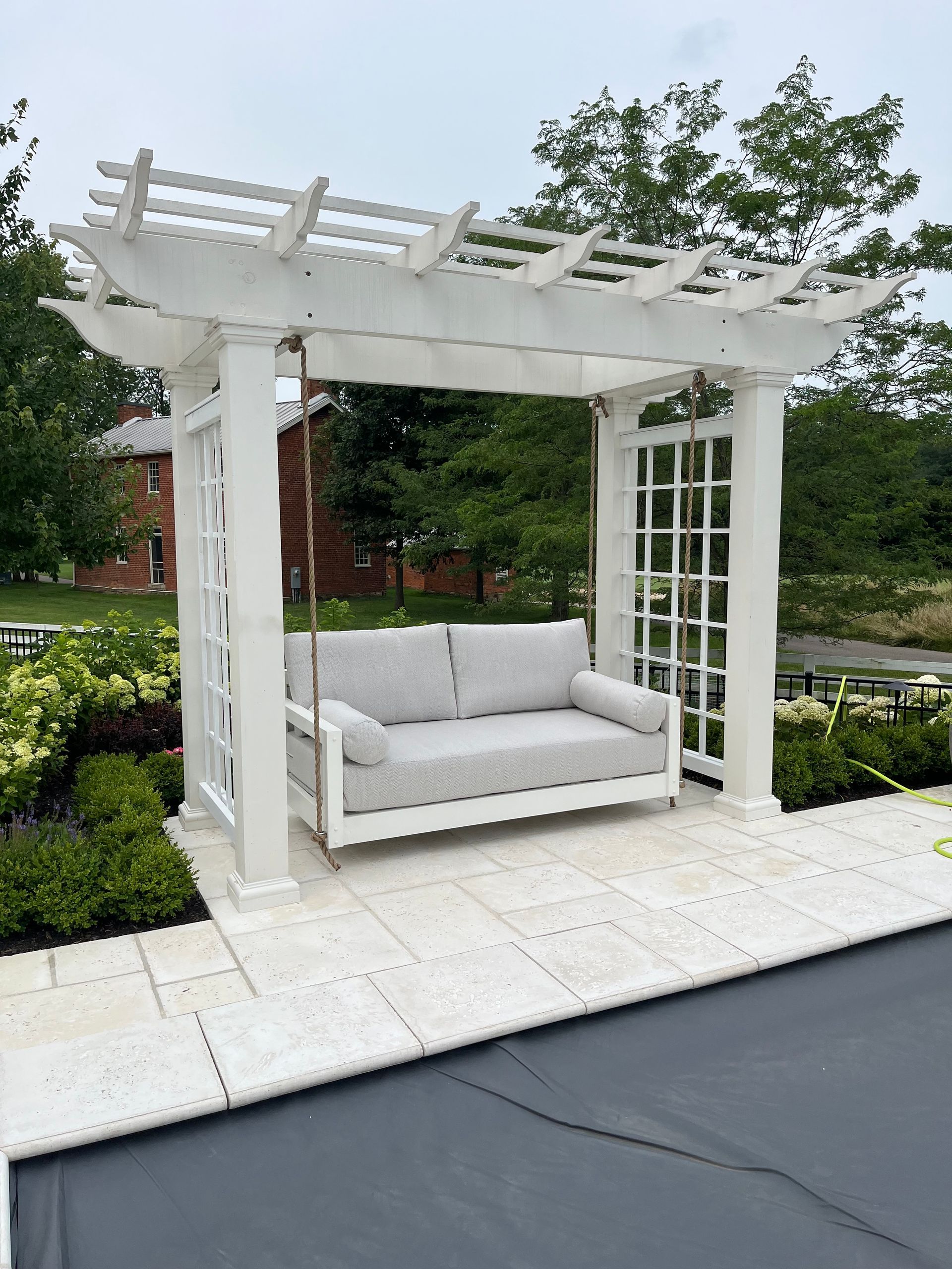 White pergola with cushioned bench swing on a patio beside a pool, surrounded by greenery.
