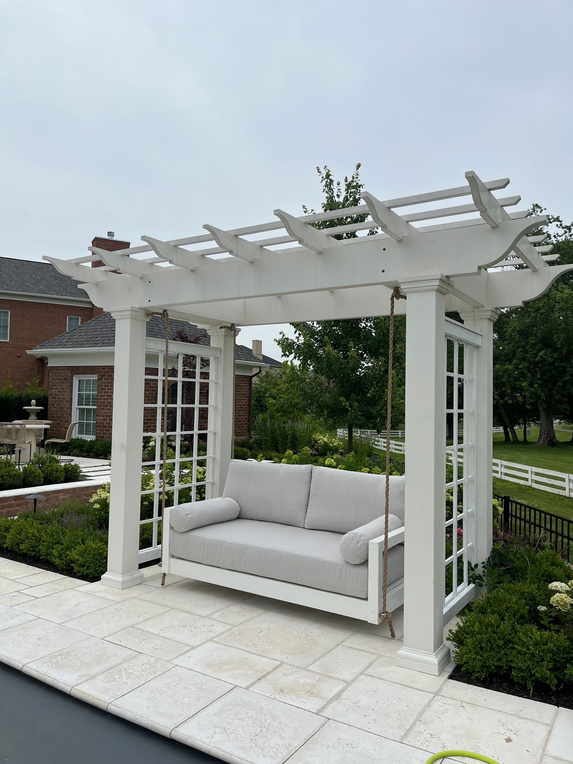 White pergola with a gray loveseat on a paved patio in a landscaped garden