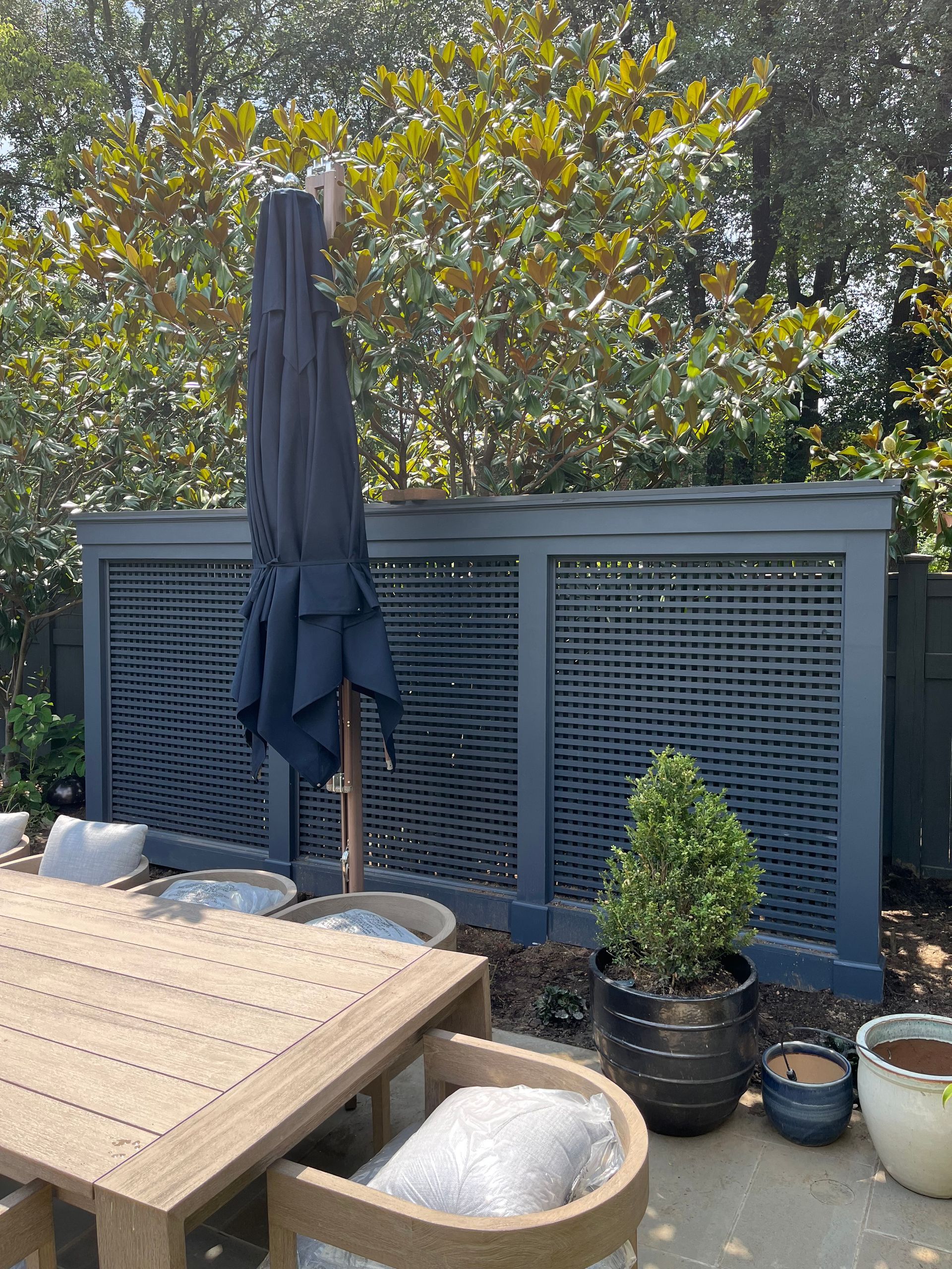 Backyard patio with covered umbrella, wooden table, black planter, and potted plants against a gray fence