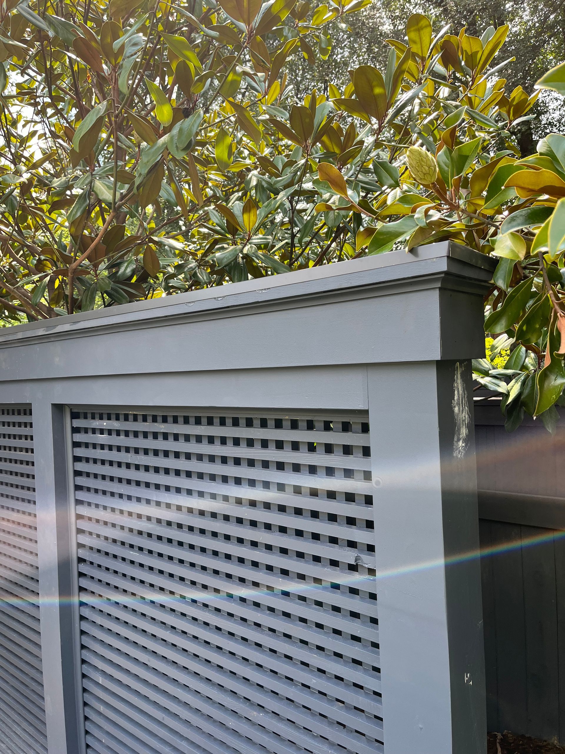 White louvered outdoor utility enclosure with gray trim in front of green foliage.