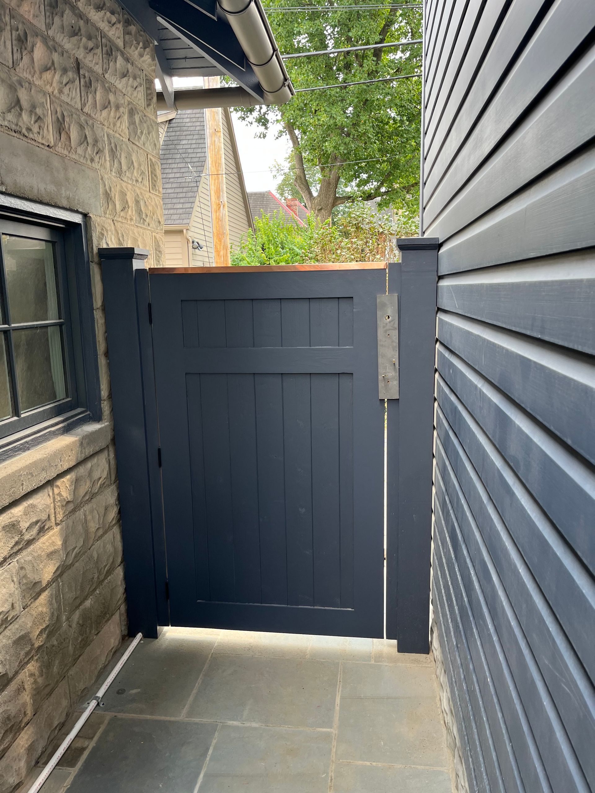 Narrow side passage with a dark blue gate between a stone wall and gray siding, leading to a garden.