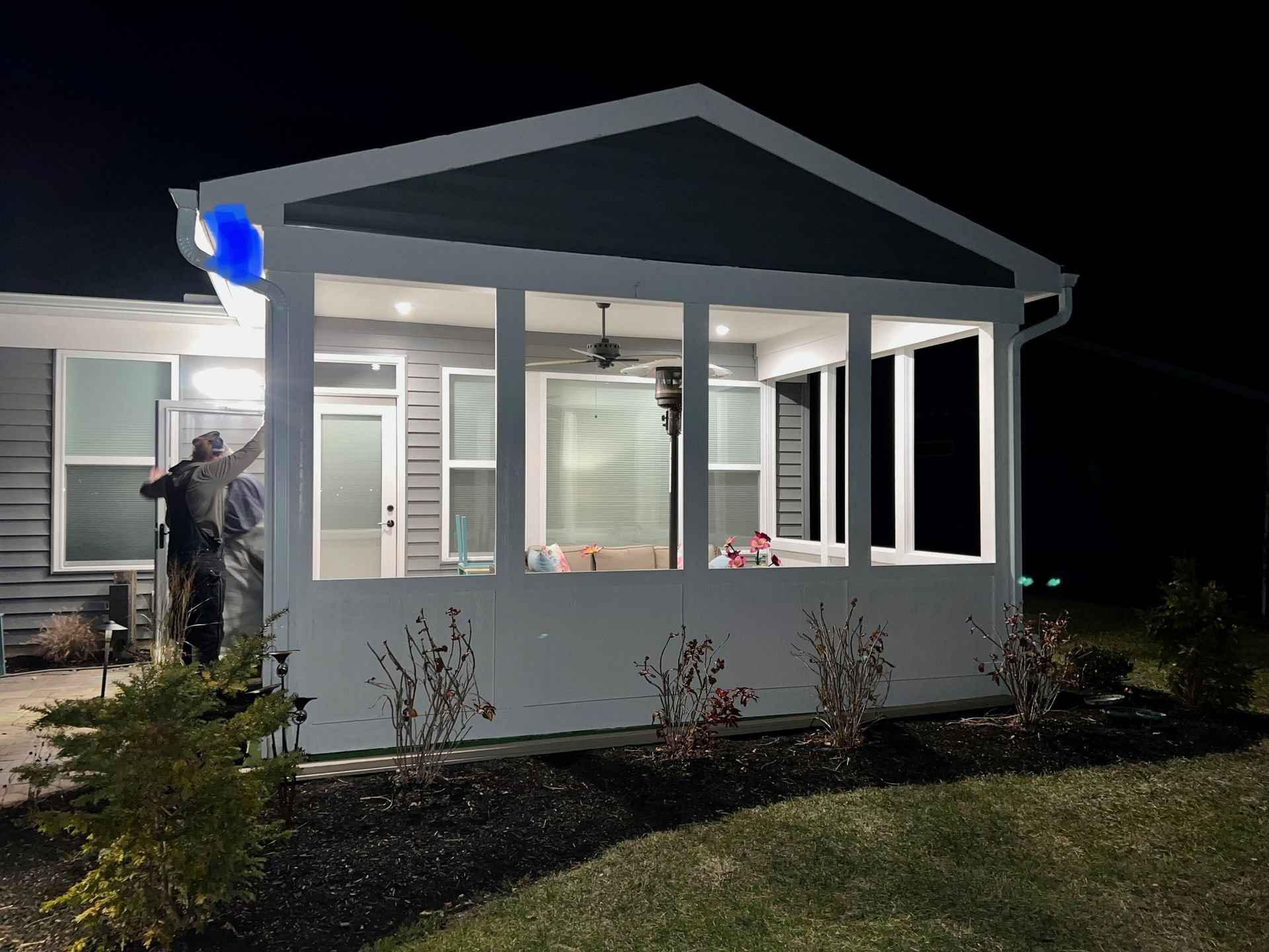 Nighttime view of a white porch addition with lit windows and a worker standing by the entrance