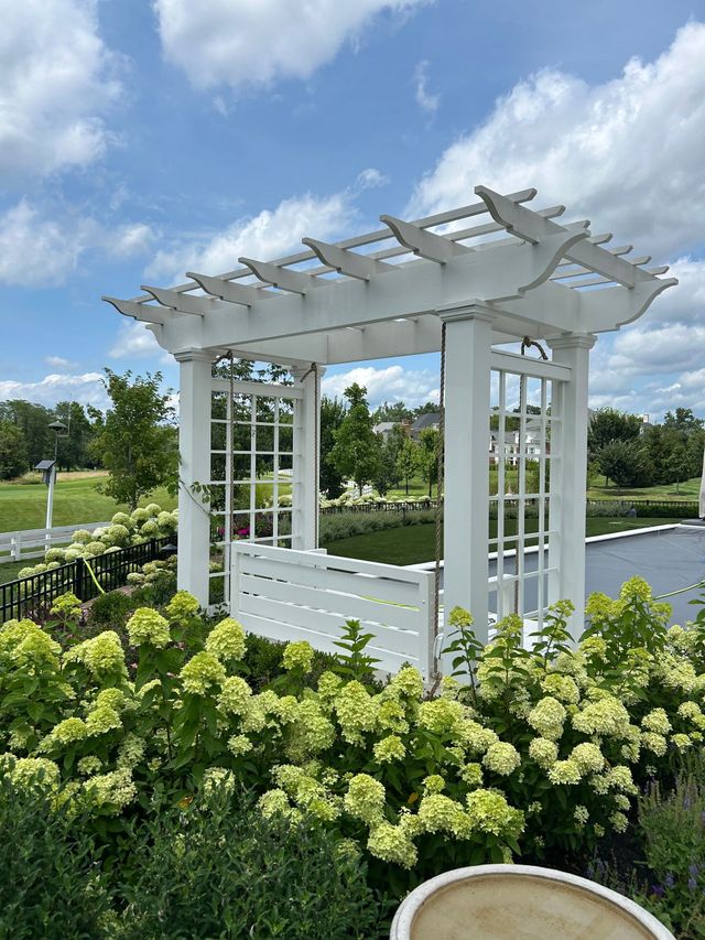 White garden arbor with trellis sides among green shrubs beside a pond under a blue sky