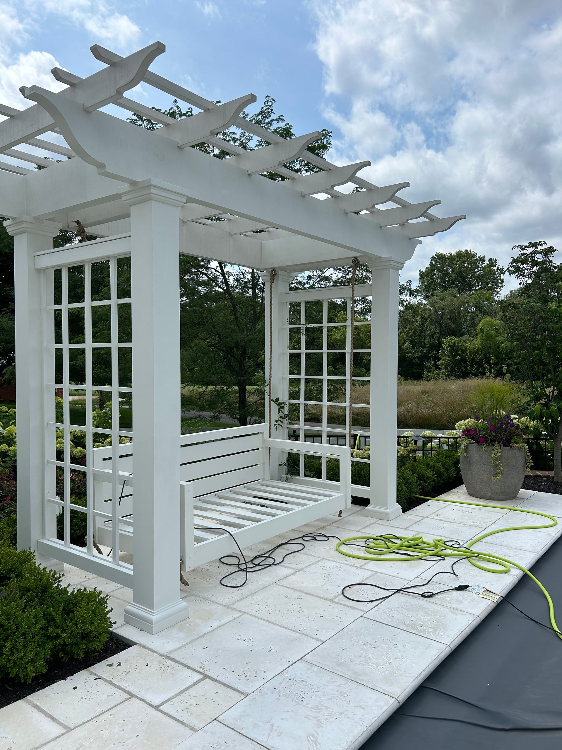 White garden pergola with swing bench on a stone patio beside greenery