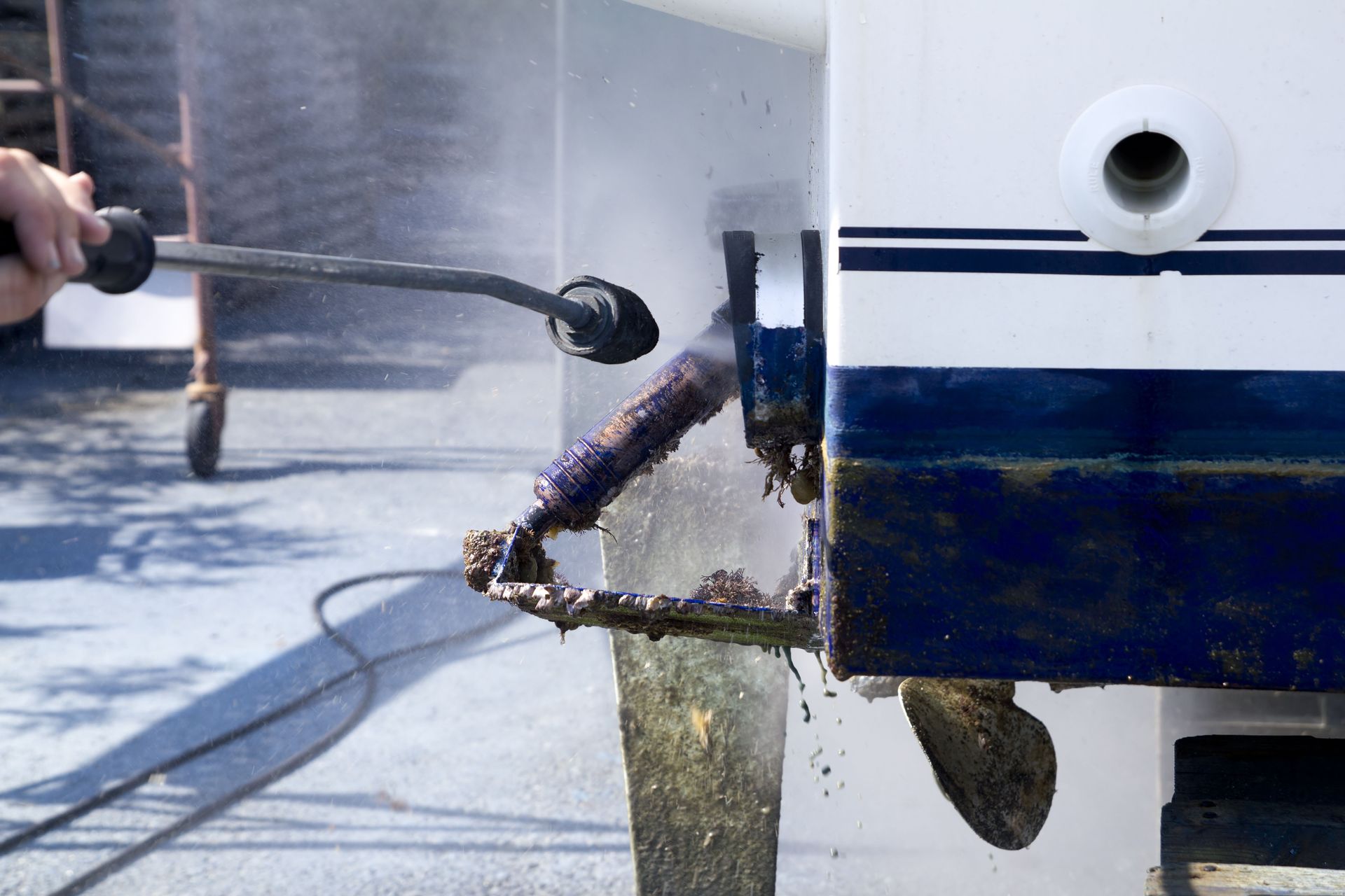 Pressure washing a boat's underwater propeller shaft, removing marine growth; blue and white boat, sunny outdoors.