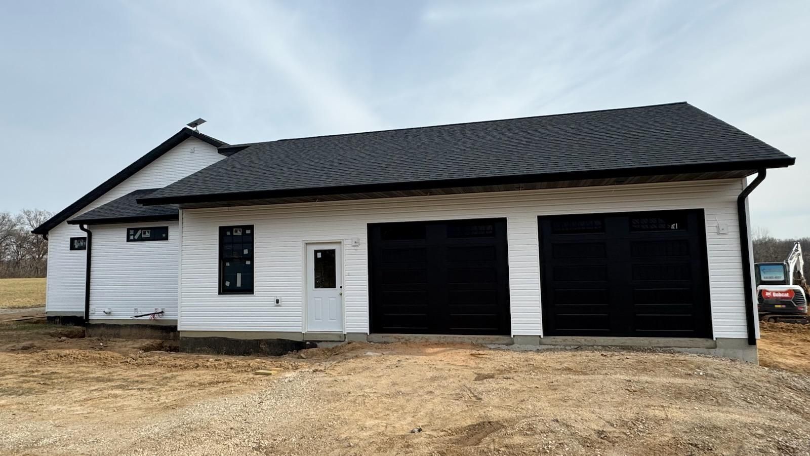 A white house with black garage doors is sitting on top of a dirt field.