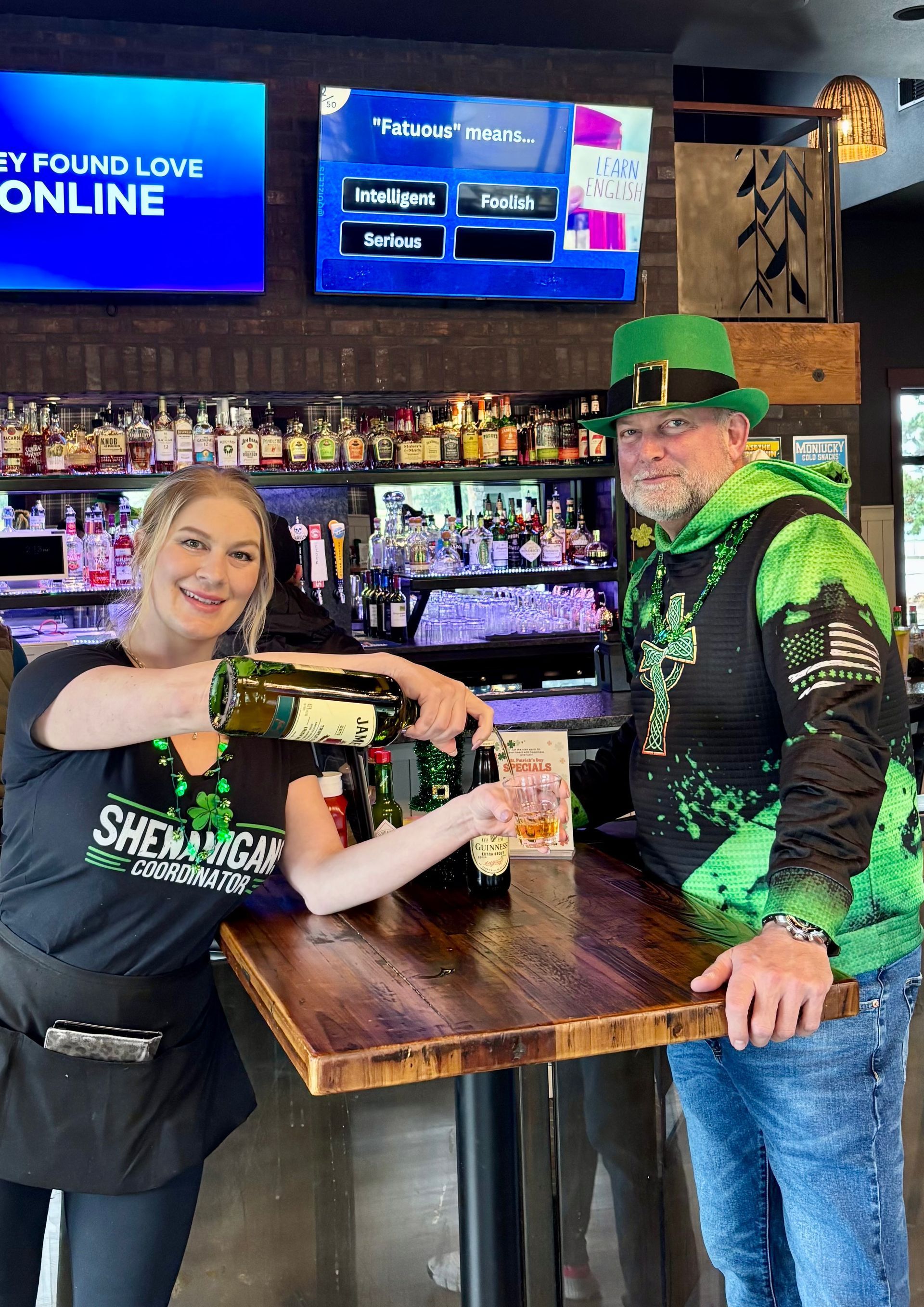 A smiling bartender pours a drink for a customer wearing a green hoodie and leprechaun hat at a bar decorated for St. Pat's.