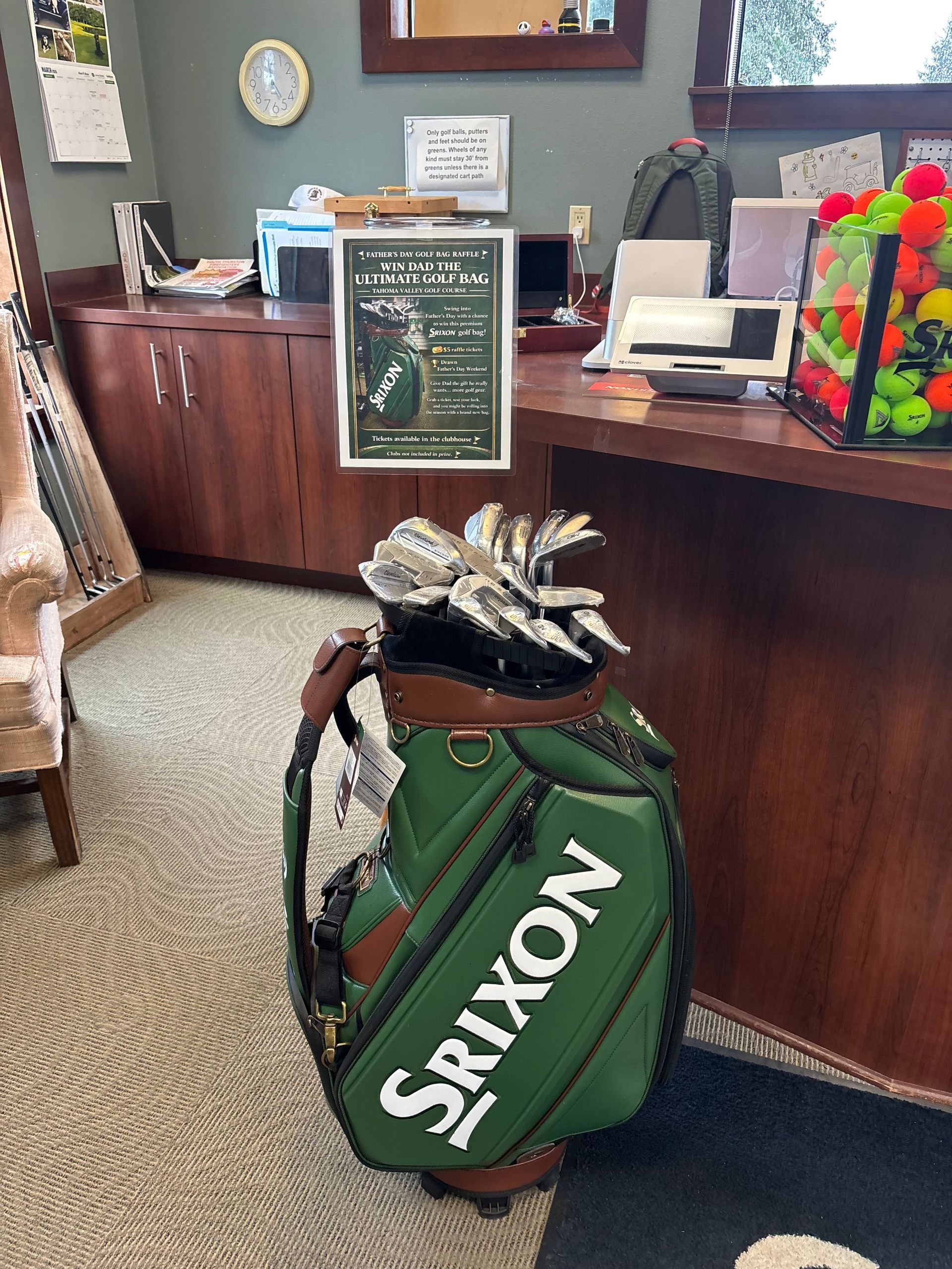 A Srixon golf bag sits on a carpeted floor in front of a wooden counter with a promotional sign and a bucket of golf balls.