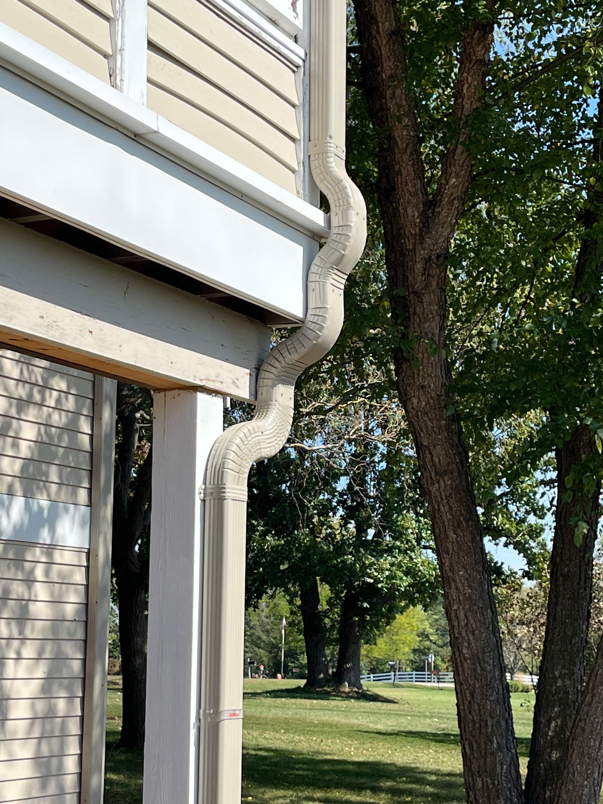 Beige zigzag downspout on a house, next to a tree, with green grass and trees in the background.