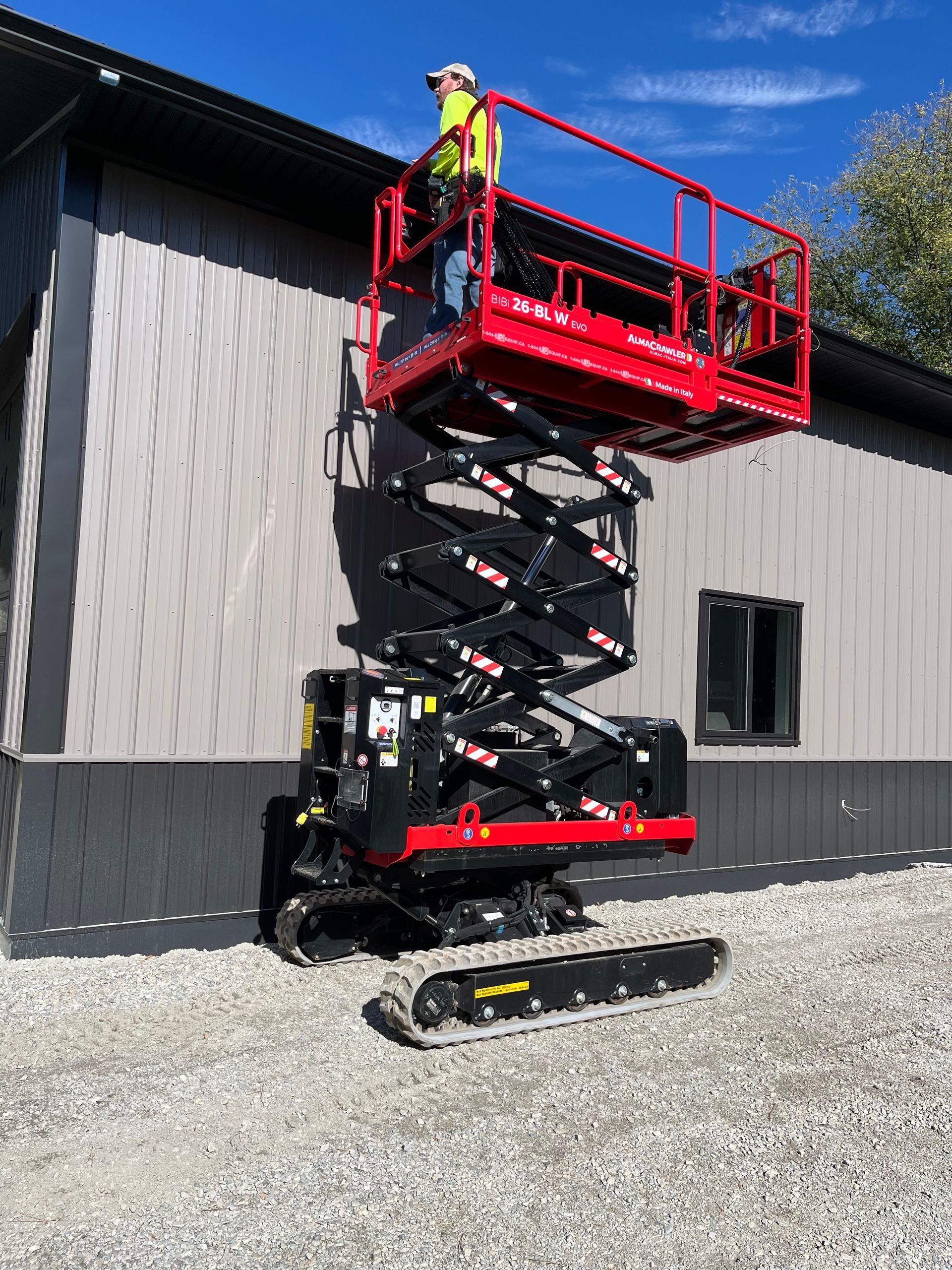 Construction worker in red scissor lift basket, working on a building exterior. Lift is on tracks, gravel ground.