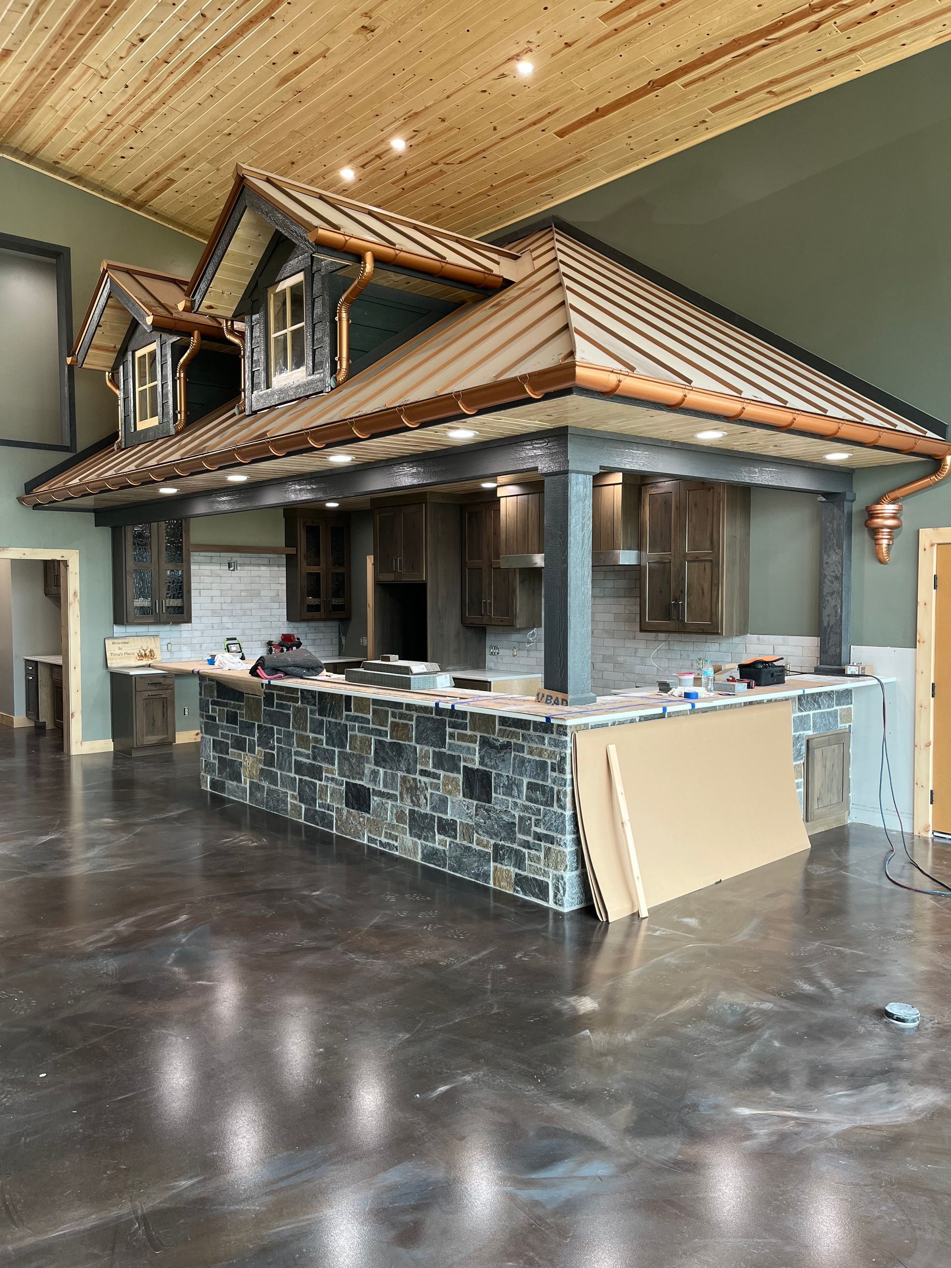 Rustic kitchen island under a decorative roof with stone facade, copper gutters, and gray cabinets.
