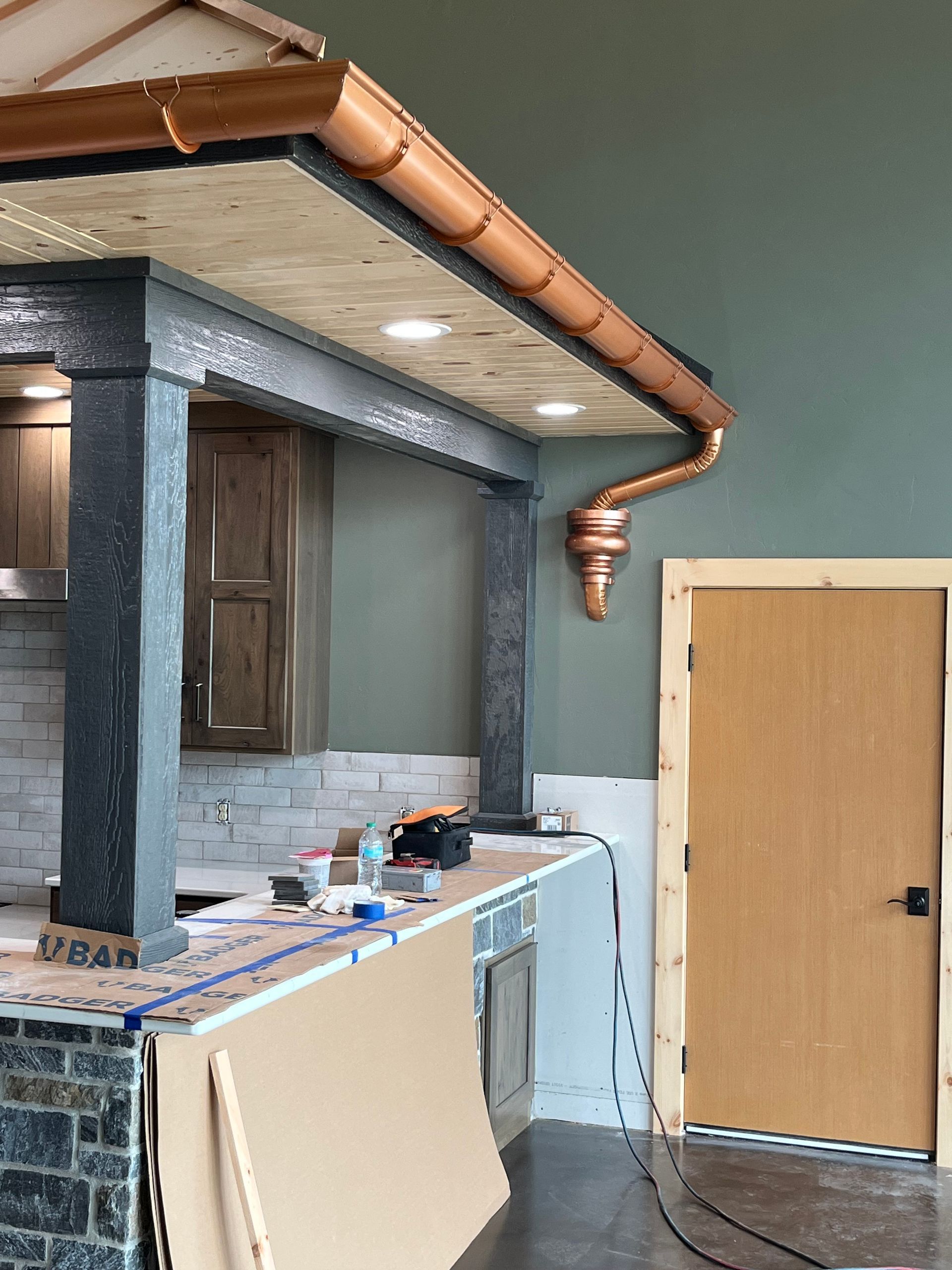 Copper gutters and downspout on a building's exterior, near a wooden door and a partially constructed bar.