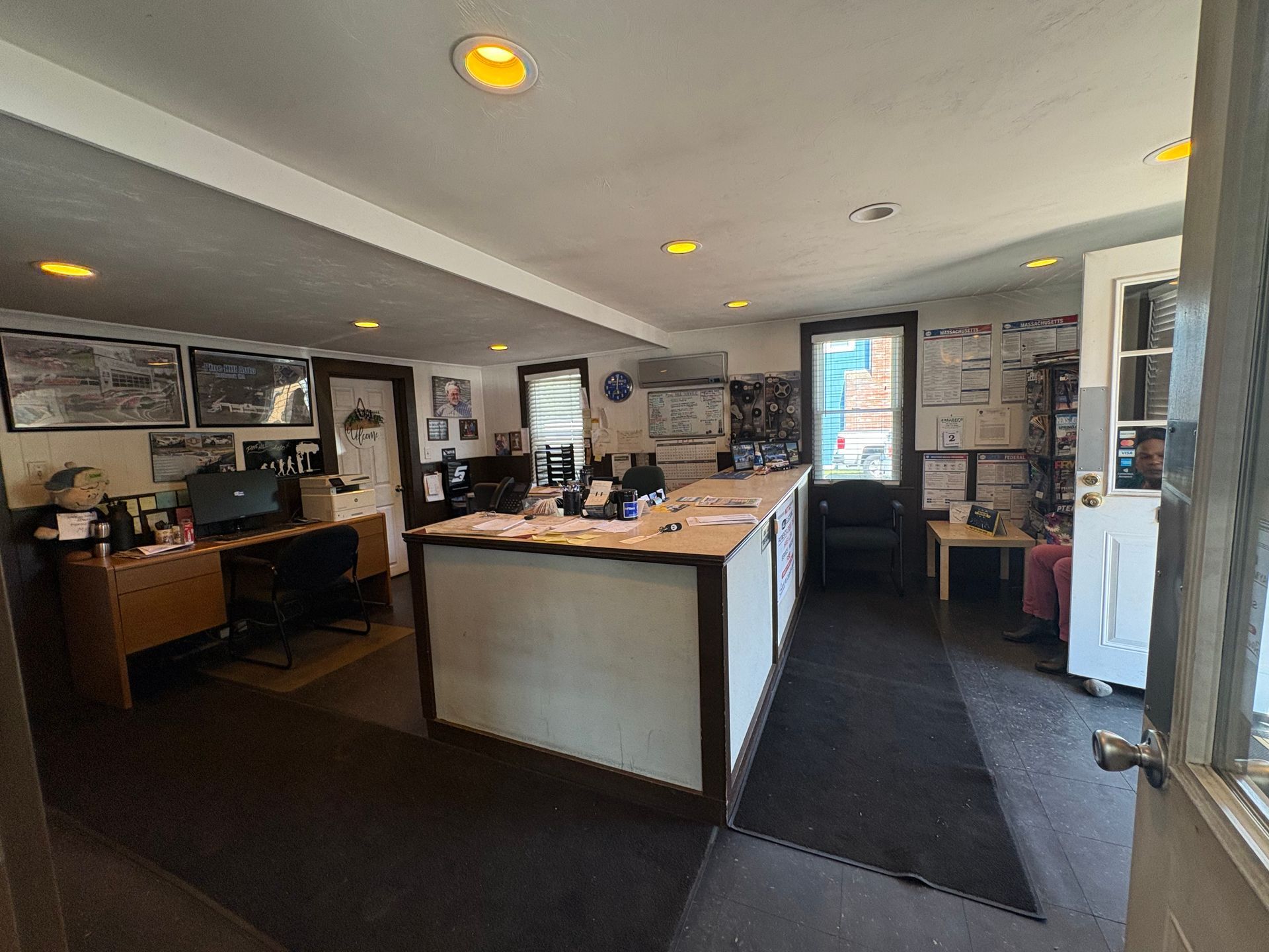 Office interior with a counter, desk, and open door. Walls feature photos and documents.