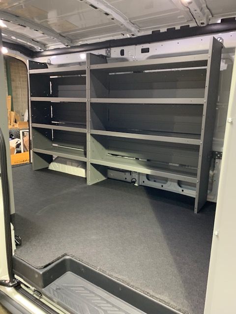Interior of a work van with gray shelving against the wall, black floor, and open doors.