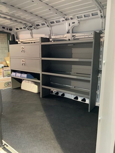 Gray shelving unit inside a white cargo van. The unit has shelves and drawers. The floor is black.