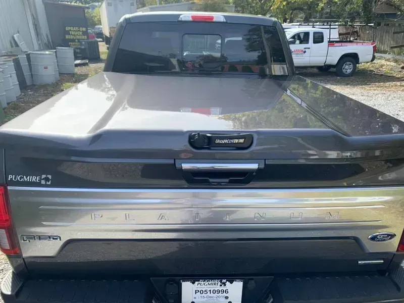 Rear view of a gray pickup truck bed with a closed cover, showing the tailgate and a white truck in the background.
