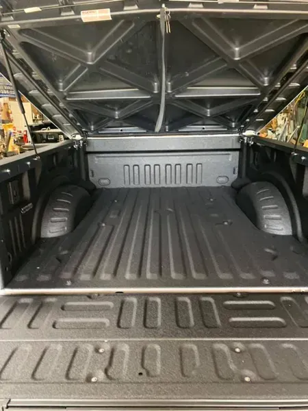 Interior of a truck bed with the cover open. Black textured bed and cover, lined floor.