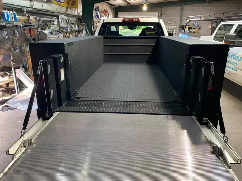 Truck bed with a retractable ramp and black storage compartments. The bed is gray and the ramp is extended.