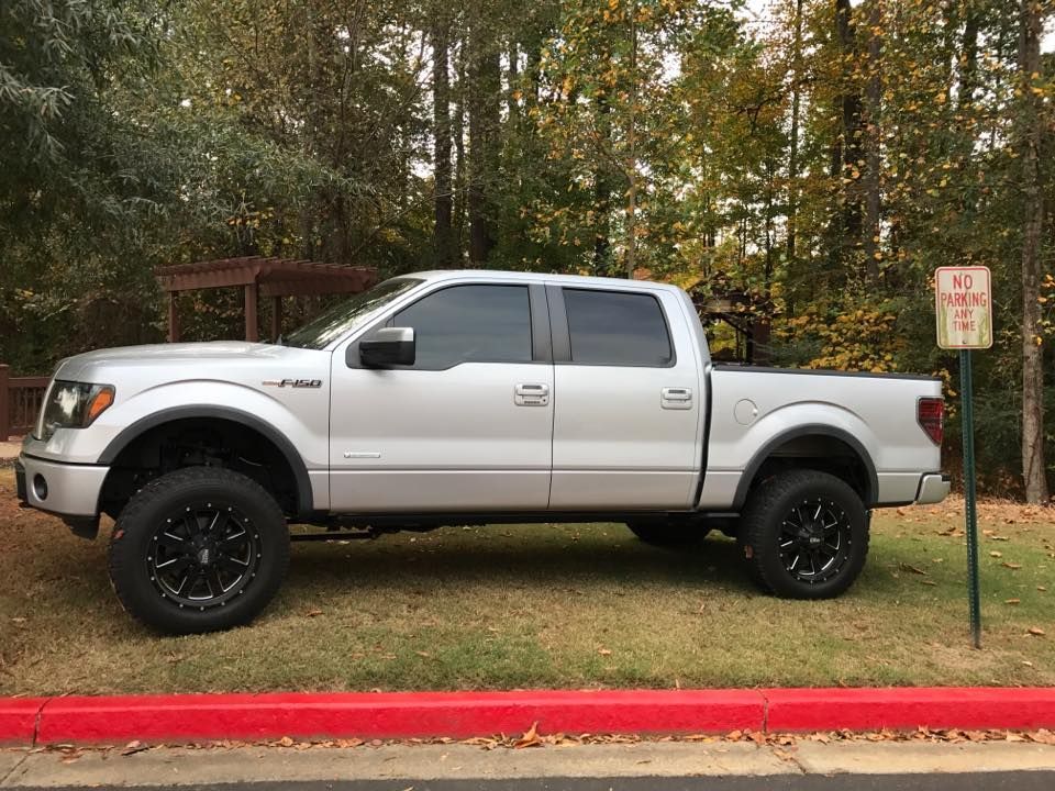 Silver Ford F-150 truck with tinted windows, black rims, parked on grass near a red curb and trees.