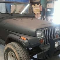 Black Jeep's front end, with a matte finish. Visible tire, headlight, and bumper. Workshop setting.
