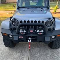 Close-up of a gray Jeep with a black bumper, tow hook, and two auxiliary lights.