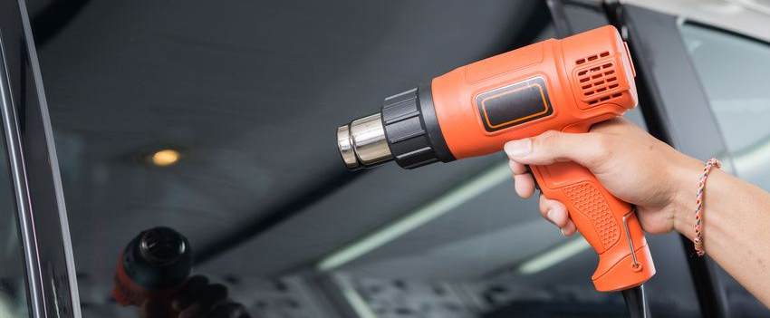 Hand holding an orange heat gun, applying heat to a car window that has tint film applied.