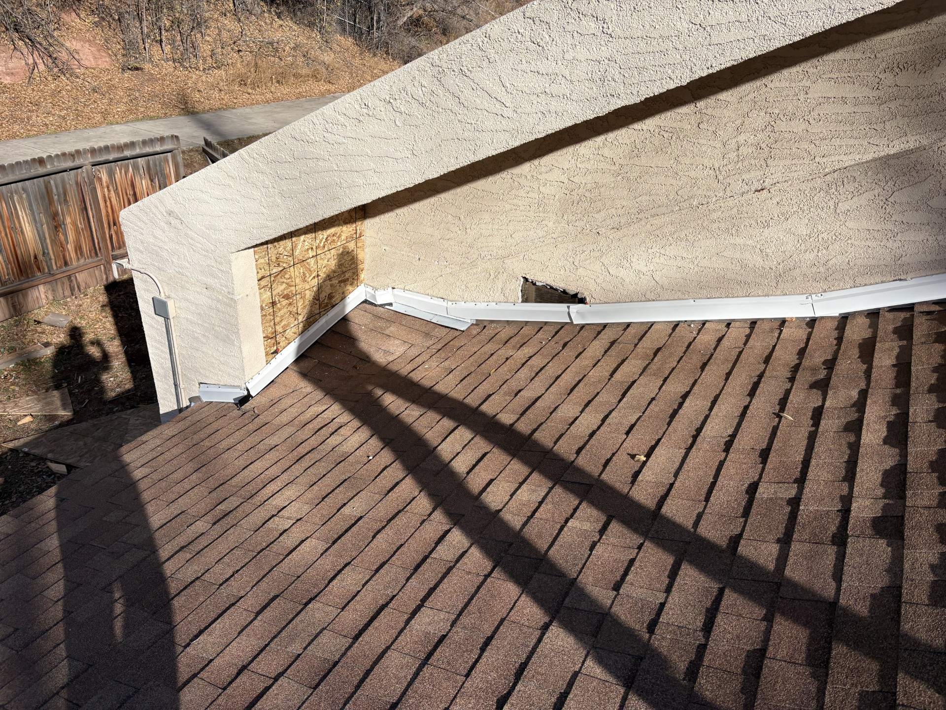 A rooftop with brown shingles and a light-colored stucco wall. Shadows stretch across the roof.