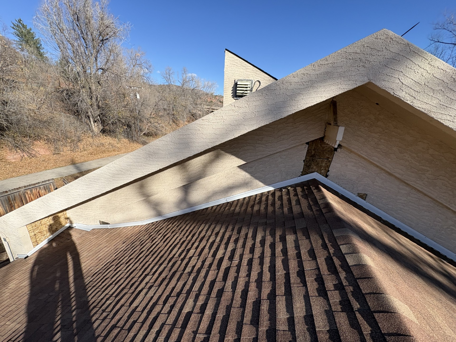 View of a building's roof intersection with brown shingles, beige walls, and a blue sky in a natural setting.