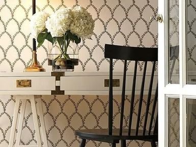 White desk with a flower arrangement and black chair against patterned wallpaper by an open door.