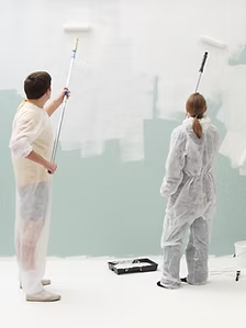 Two people painting a wall with rollers; one wears a coverall and the other a protective apron.