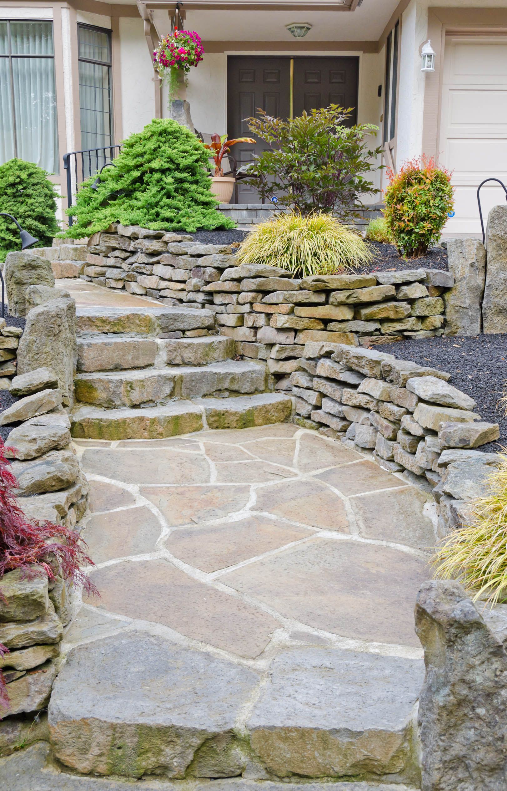 Stone front walkway with steps leading to a house entrance, bordered by low retaining walls and plants