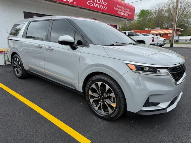 Silver minivan parked in a lot, front-left view, with yellow parking lines and a red store sign in the background.